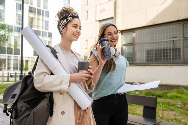 Two women female students walking together in front of university building in summer day holding notes and talking discuss about exam or lesson real people caucasian female colleagues copy space