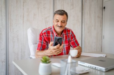 Üst düzey bir erkek girişimci iş yerindeki masada oturuyor bir fincan kahve ve cep telefonu tutuyor online mesajları veya sosyal medya içeriğini kontrol ediyor gerçek insanlar boşluğu kopyalıyor