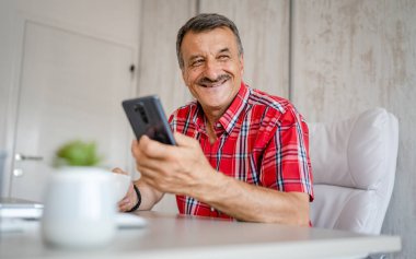 one senior man male entrepreneur sitting at the desk table at office at work taking a brake hold cup of coffee and mobile phone checking online messages or social media content real people copy space