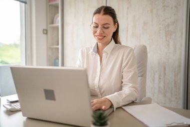 Beyaz gömlekli, beyaz gömlekli, iş kadını ya da sekreterli bir kadın bilgisayarının masasında oturuyor, gülümsüyor.