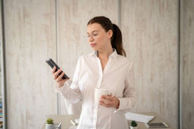 One woman caucasian female businesswoman entrepreneur manager or secretary standing in her office front view copy space happy smile confident with cup of coffee using mobile phone to check messages