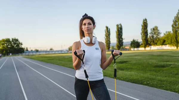 One woman adult caucasian female athlete training with elastic rubber resistance bands in stadium on track in summer evening or morning happy brunette health and fitness concept copy space