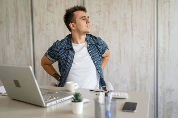 One man freelancer stretch while sitting in the office at work sitting in chair at desk using laptop computer having pain sore back suffering from neck ache overwork and exhaustion concept copy space