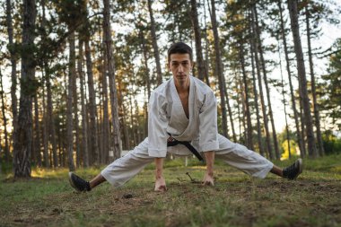 One man caucasian karateka male athlete training karate in the forest in woods in nature wear kimono gi uniform real people copy space martial arts concept stretching