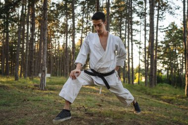 One man caucasian karateka male athlete training karate in the forest in woods in nature wear kimono gi uniform real people copy space martial arts concept stretching