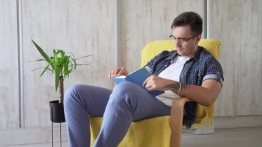 One man caucasian male young adult sitting in a chair at home reading a book wear blue shirt and wear eyeglasses leisure time concept education copy space