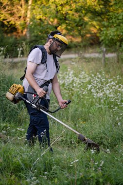 Young caucasian man farmer gardener standing in the field with string trimmer petrol Brushcutter cut weed grass working on farm cutting in the field in sunny day in the village side view