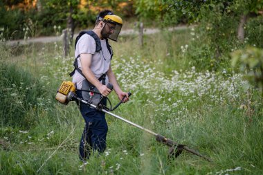 Young caucasian man farmer gardener standing in the field with string trimmer petrol Brushcutter cut weed grass working on farm cutting in the field in sunny day in the village side view