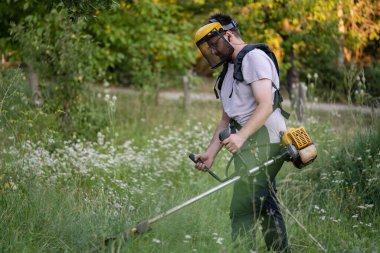 Young caucasian man farmer gardener standing in the field with string trimmer petrol Brushcutter cut weed grass working on farm cutting in the field in sunny day in the village side view