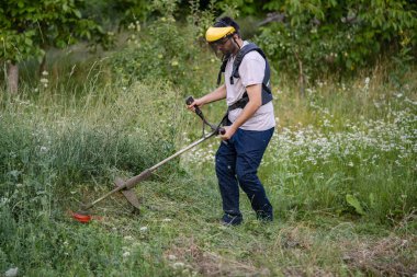 Young caucasian man farmer gardener standing in the field with string trimmer petrol Brushcutter cut weed grass working on farm cutting in the field in sunny day in the village side view