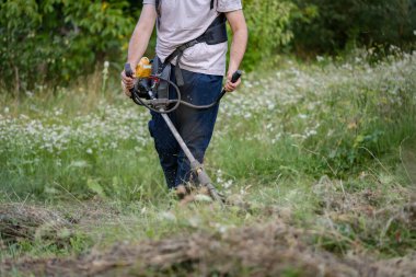 Young caucasian man farmer gardener standing in the field with string trimmer petrol Brushcutter cut weed grass working on farm cutting in the field in sunny day in the village midsection