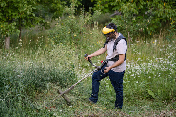 Молодой кавказский фермер-садовник, стоящий в поле с бензиновым триммером, подстригал траву, работая на ферме в поле в солнечный день в сельской местности.