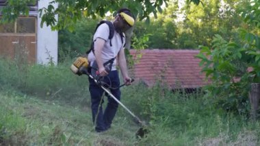 Young caucasian man farmer gardener standing in the field with string trimmer petrol Brushcutter cut weed grass working on farm cutting in the field in sunny day by the house in the village