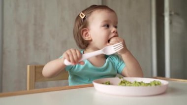 one girl small caucasian child toddler sitting at the table at home eating food alone using plastic fork childhood growing up and development concept copy space healthy eating 