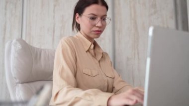 One woman female caucasian entrepreneur businesswoman or secretary sitting at her office at desk work on laptop computer typing happy smile wear shirt and eyeglasses making phone call
