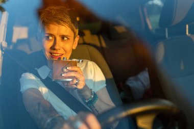 One woman mature caucasian female with tattoo on arms hold flask while sitting in the car alone driver on parked automobile drinking alcohol abuse and alcoholism concept copy space front view