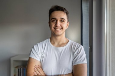 One man young adult male caucasian with brown hair standing by the window at home in day confident looking to the camera happy smile wear white shirt real people copy space