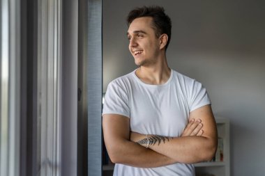 One man young adult male caucasian with brown hair standing by the window at home in day confident looking to the side happy smile wear white shirt real people copy space