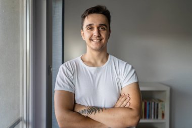 One man young adult male caucasian with brown hair standing by the window at home in day confident looking to the camera happy smile wear white shirt real people copy space