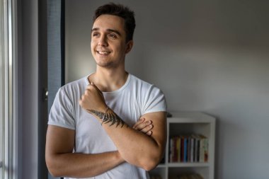 One man young adult male caucasian with brown hair standing by the window at home in day confident looking to the side happy smile wear white shirt real people copy space