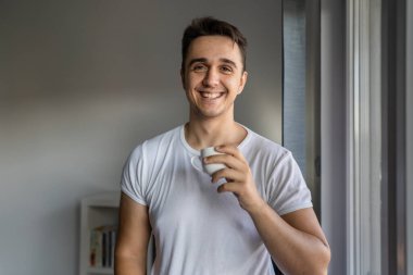 One man young adult male caucasian with brown hair standing by the window at home in morning day confident looking to the camera happy smile confident wear white shirt with cup of coffee copy space