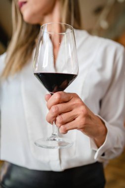 close up on midsection of unknown caucasian woman hold glass of red wine while standing indoor wear white shirt copy space