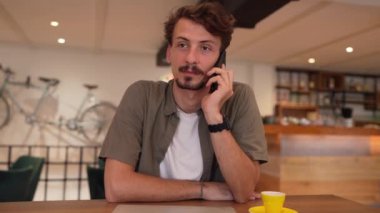 one man young adult millennial caucasian male sitting at the table alone in cafe using mobile phone making a call talking happy confident front view wear casual shirt with mustaches and beard modern