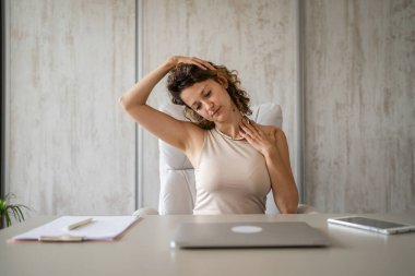 Ofiste çalışan serbest çalışan bir kadın dizüstü bilgisayarla masa başında oturuyor. Boyun ağrısı, aşırı çalışma ve yorgunluk yüzünden sırtı ağrıyormuş.