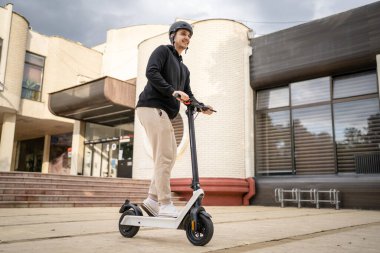 Genç, yetişkin, beyaz bir erkek. Elektrikli scooterın üzerinde duruyor. Şehirde ya da şehirde araba sürüyor.