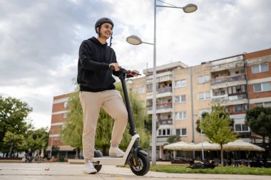 Genç, yetişkin, beyaz bir erkek. Elektrikli scooterın üzerinde duruyor. Şehirde ya da şehirde araba sürüyor.