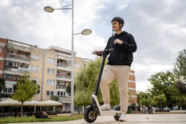 Genç, yetişkin, beyaz bir erkek. Elektrikli scooterın üzerinde duruyor. Şehirde ya da şehirde araba sürüyor.