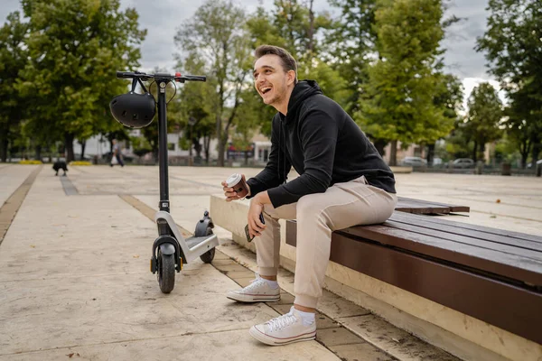 一人の若いです大人Cucasian男性座っていますザベンチで市内や町によって電動キックスクーター電子スクーターで一日待っています本当の人々コピースペース幸せな笑顔 — ストック写真