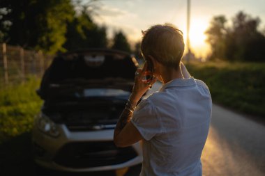 Olgun bir kadın akşamüstü yolda duruyor. Bozuk araba otomobili kaputu açık. Yol yardımı çekme konsepti için telefon ediyor.