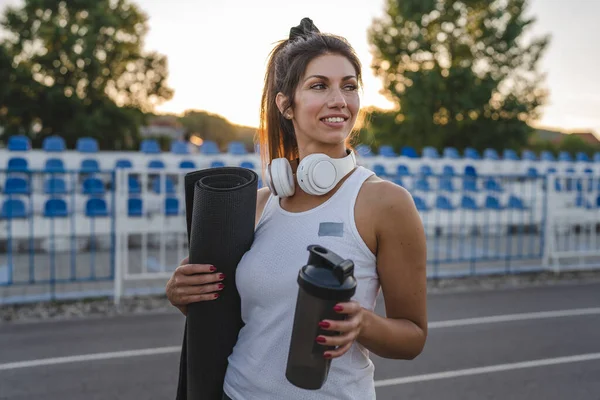 one woman adult caucasian female athlete standing on stadium on running track with supplement shaker and yoga mat with headphones ready for training outdoor in summer evening real people copy space