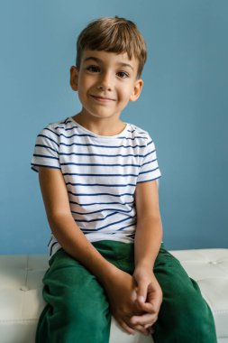 Anaokulundan beyaz bir çocuk evdeki kanepede oturmuş mavi duvarın önünde oturmuş gülümsüyor.
