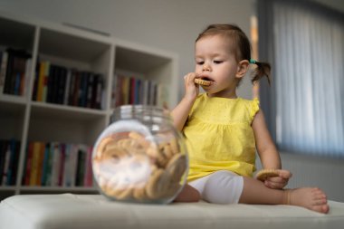 Bir küçük beyaz bebek evde oturup cam kavanozdan kurabiye yiyor ve çocukluk konseptini evde kopyalıyor.
