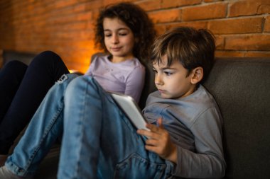Küçük beyaz çocuk evde dijital tablet kullanarak kız kardeşinin yanında oturuyor.