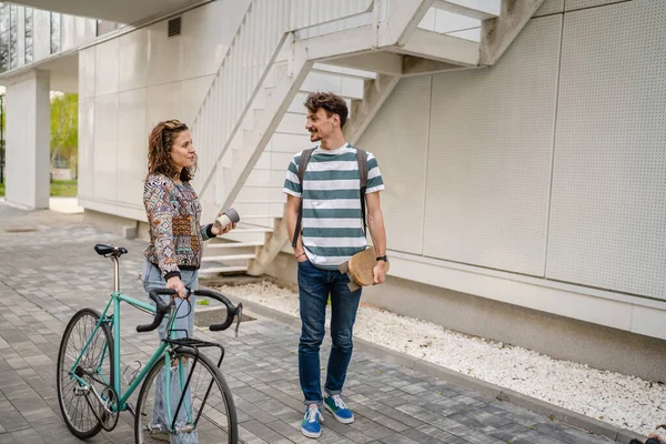 Two people young adult couple man and woman boyfriend and girlfriend or students friends walk by the building or university in day with bicycle and skateboard modern lifestyle concept copy pace