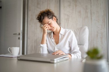 Ofisteki masada oturan bir kadın aşırı yorgun ve bitkin bir halde dizüstü bilgisayarın uzun kullanımından dolayı göz yorgunluğu çekiyor.
