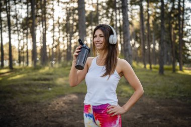 One woman happy caucasian female standing in the forest or park in woods with supplement shaker and headphones during training in nature taking a brake water drink and hydration real people copy space