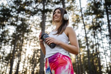 One woman happy caucasian female standing in the forest or park in woods with supplement shaker and headphones during training in nature taking a brake water drink and hydration real people copy space