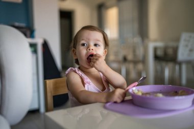 Bir çocuk, küçük beyaz bir bebek, evde masada tek başına yemek yiyor. Gerçek insanlar, erken dönem çocuk gelişimi konseptini taklit ediyor.