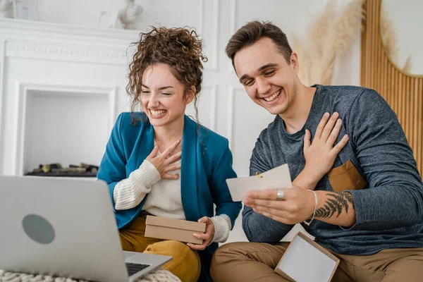 Young caucasian couple woman and man wife and husband or girlfriend and boyfriend opening gifts box presents and read card in front of laptop computer at home having online video call copy space
