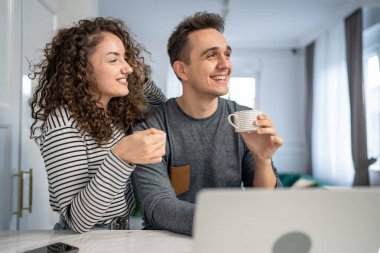 İki yetişkin genç çift karı koca ya da karı koca ya da kız arkadaş mutfakta dizüstü bilgisayarla çevrimiçi içerik alışverişini kontrol ediyorlar ya da video sabah rutini yapıyorlar.