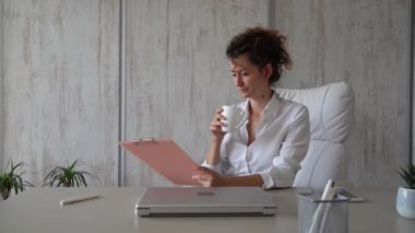 Ofiste çalışan beyaz bir kadın. Elinde bir pano var ve kahve içiyor. Modern iş kadını konsepti.