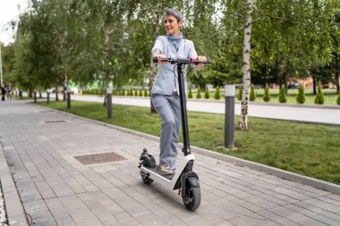 Yetişkin, yaşlı, beyaz bir kadın. Elektrikli scooterın üzerinde duruyor. Şehirde ya da şehirde araba sürüyor.