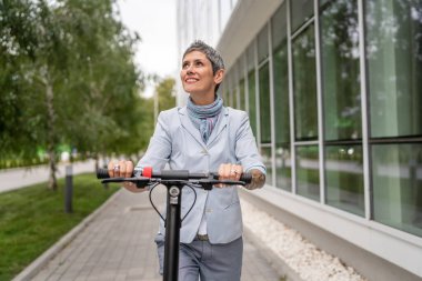 Yetişkin bir son sınıf beyaz kadın elektrik tekmeli scooter 'a biniyor. Şehirde ya da şehirde araba sürüyor. E-scooter çevre dostu.