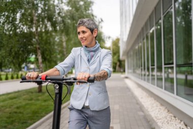 Yetişkin bir son sınıf beyaz kadın elektrik tekmeli scooter 'a biniyor. Şehirde ya da şehirde araba sürüyor. E-scooter çevre dostu.