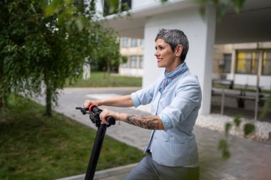 Yetişkin bir son sınıf beyaz kadın elektrik tekmeli scooter 'a biniyor. Şehirde ya da şehirde araba sürüyor. E-scooter çevre dostu.