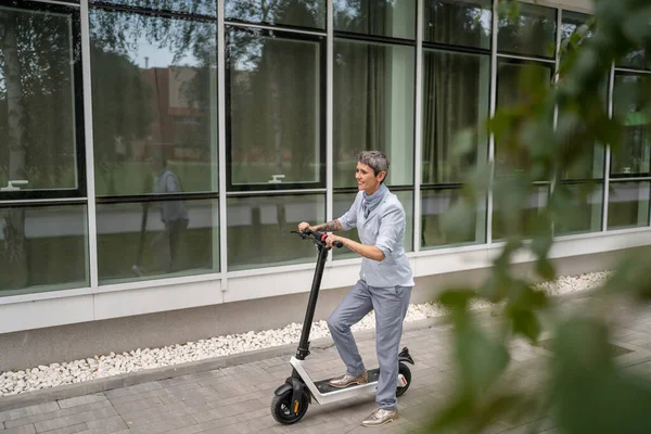 one woman mature senior caucasian female standing on electric kick scooter in day riding driving in town or city e-scooter eco friendly mode of transport real people copy space full length